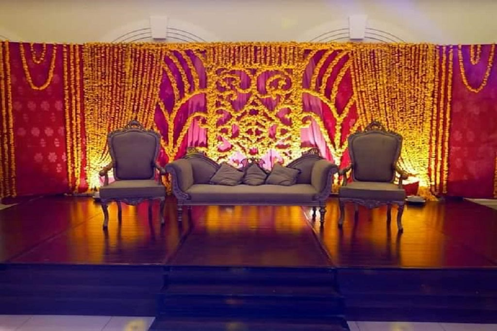 A wedding setup with a yellow and pink floral backdrop, marigold flower decorations, and elegant seating for the bride and groom in a vibrant, festive setting.