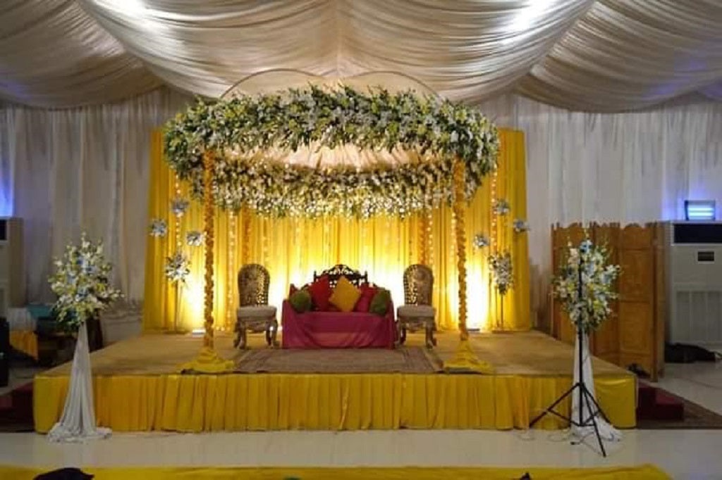 A wedding mandap setup with a floral canopy in yellow and white, golden pillars, colorful seating with cushions, and a vibrant yellow backdrop for a traditional wedding ceremony.