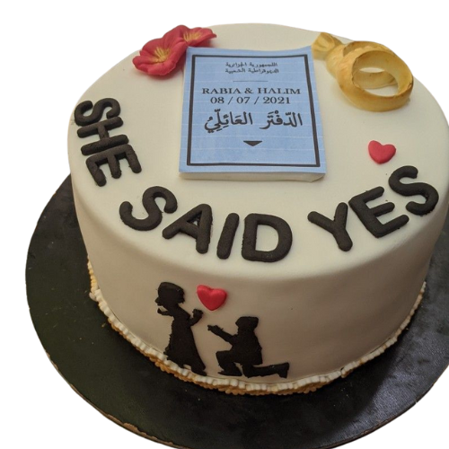 Engagement cake with She Said Yes message, wedding rings, floral accents, and proposal silhouette.