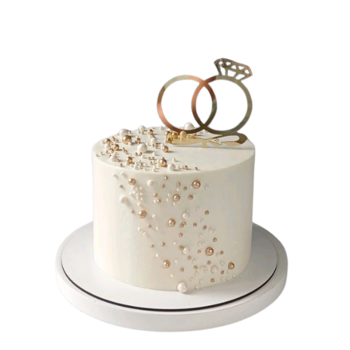 Engagement cake with white icing, pearl decorations, and golden wedding rings topper.