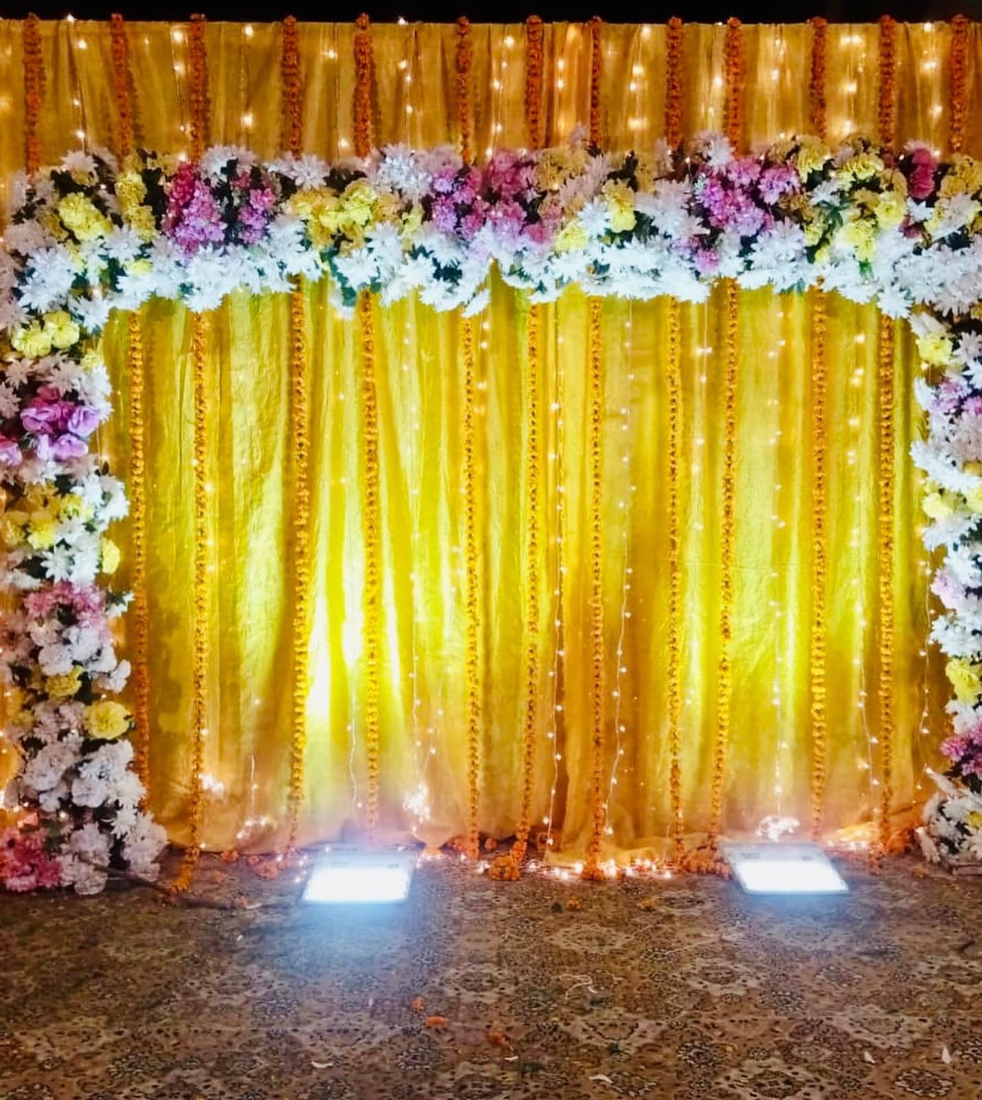 A vibrant floral wedding backdrop featuring yellow drapes, pink, white, and yellow flowers, with soft glowing string lights, perfect for an Indian wedding or mehendi ceremony.