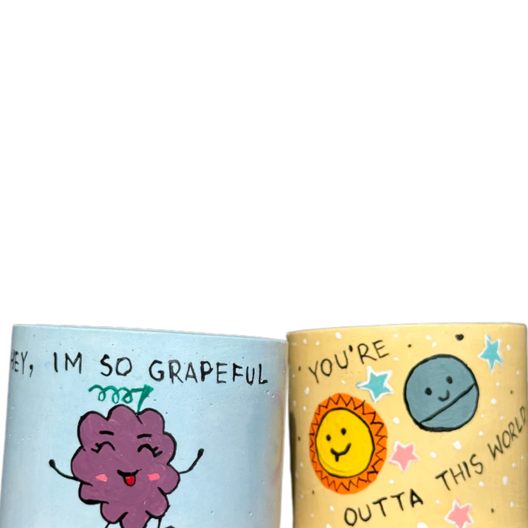 Set of two 5-inch cylindrical Painted Planters with Haworthia succulents, featuring green leaves with white spots, in yellow and blue pots with hand-painted designs of a sun, planet, and grapes, with messages “You’re Outta This World” and “Hey, I’m So Grapeful For You.”