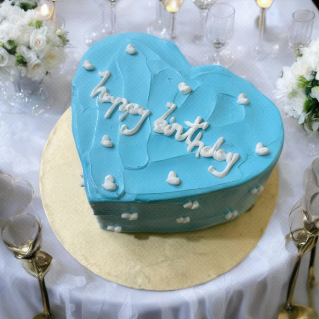 Heart-shaped blue birthday cake with white lettering, on gold board, surrounded by glasses and flowers.