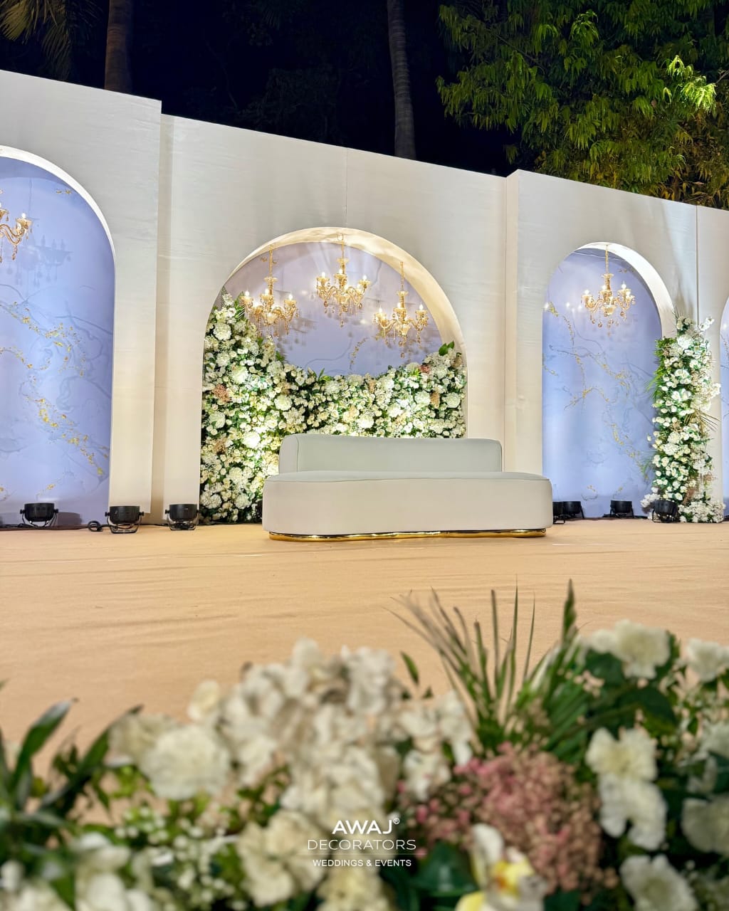 Luxury wedding stage with white sofa, floral arches, and golden chandeliers.