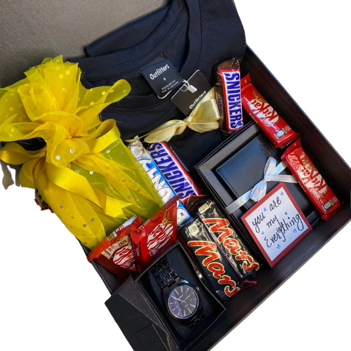 Luxury men’s gift hamper with Outfitters black t-shirt, wristwatch, leather wallet, assorted chocolates (Snickers, KitKat, Mars), yellow-wrapped gift, and mini greeting card.