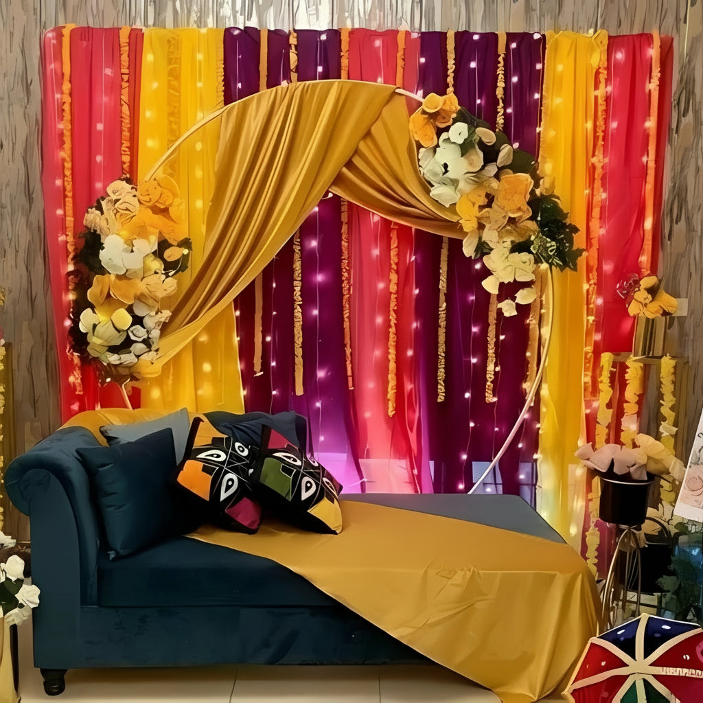Mehndi decor with a circular floral frame, yellow and white flowers, colorful drapes, fairy lights, and a modern blue sofa with vibrant cushions.