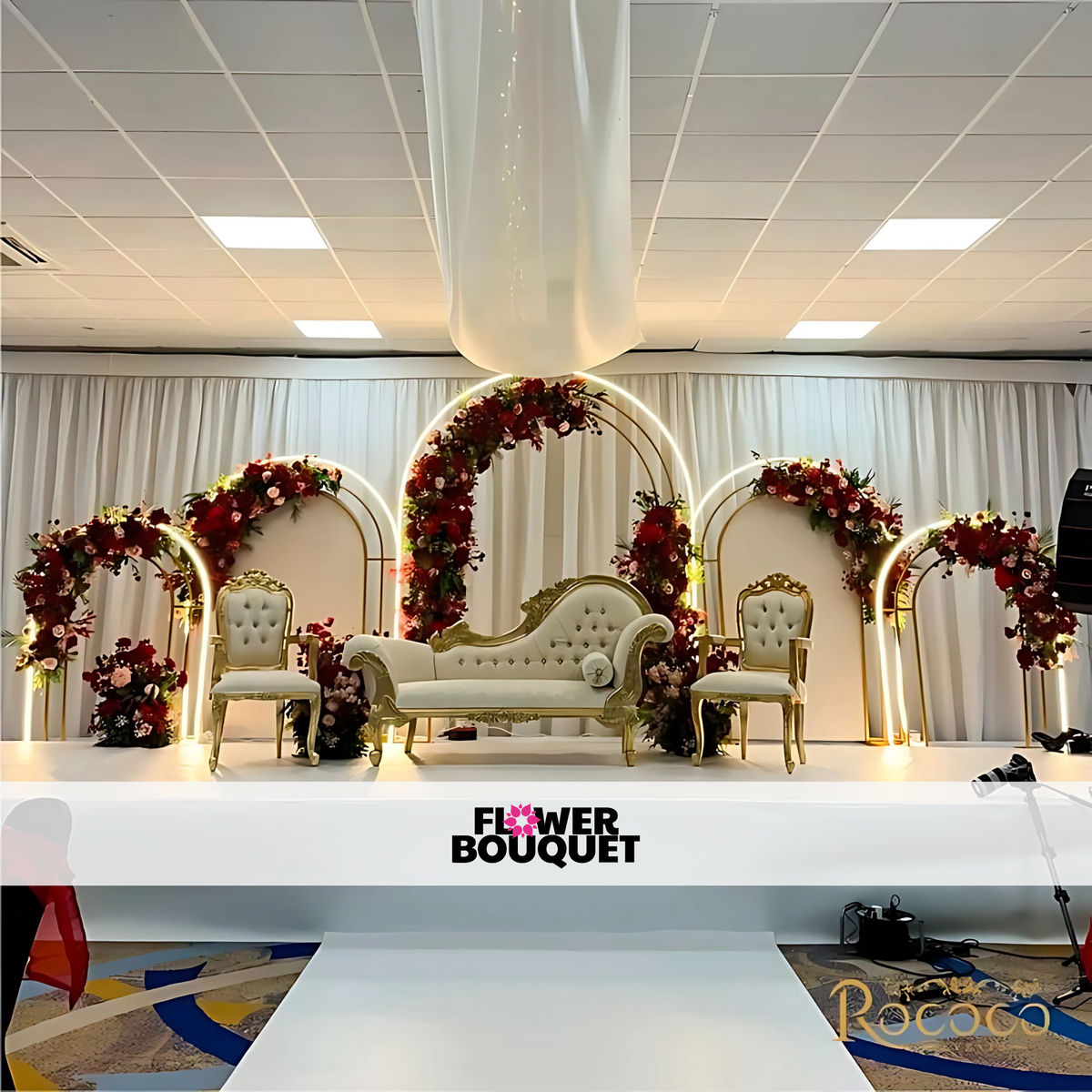 Modern wedding stage featuring golden arches with red and blush floral arrangements, a white and gold sofa, and matching chairs, ideal for luxurious celebrations.
