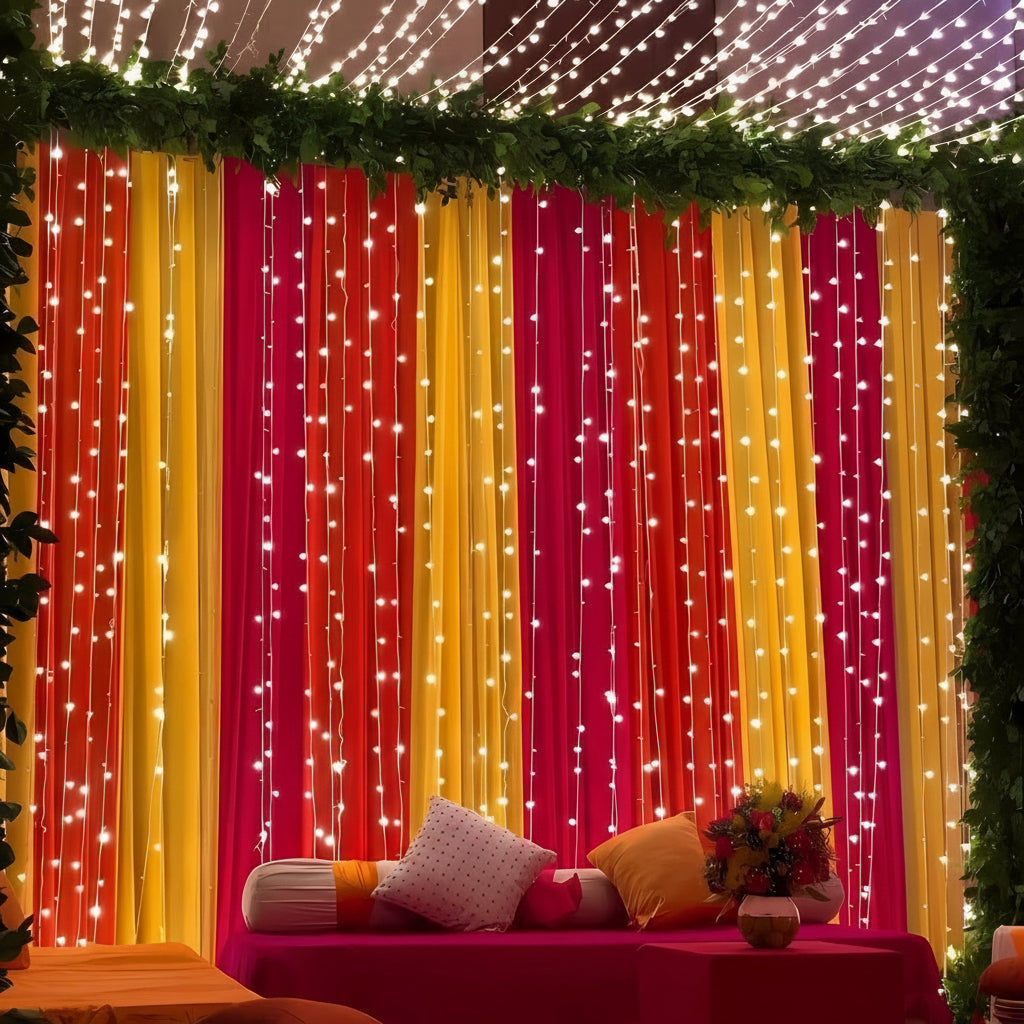 Mehndi decor with red, yellow, and pink drapes, fairy lights, and lush greenery, creating a vibrant and festive backdrop for celebrations.