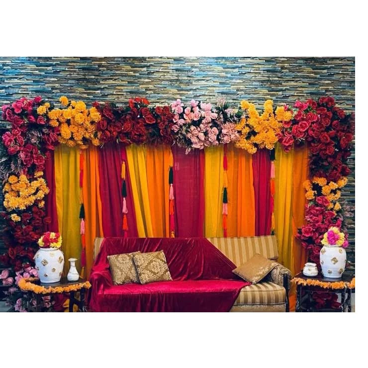 A vibrant wedding backdrop with marigold and rose flower arrangements, pink, yellow, and red fabric drapes, and a luxurious seating area adorned with traditional decorative elements for a wedding ceremony.