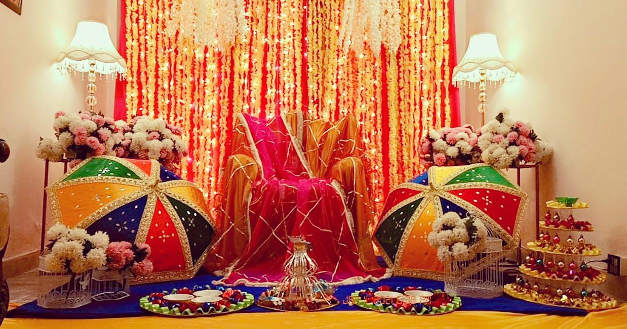 Bright wedding backdrop with marigold flowers, colorful umbrellas, floral decorations, a seating area with pink and orange hues, and twinkling lights for an event setup.