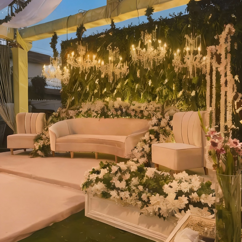 Wedding stage featuring a green backdrop with white floral arrangements, crystal chandeliers, a white curved sofa, and matching chairs, perfect for outdoor events.